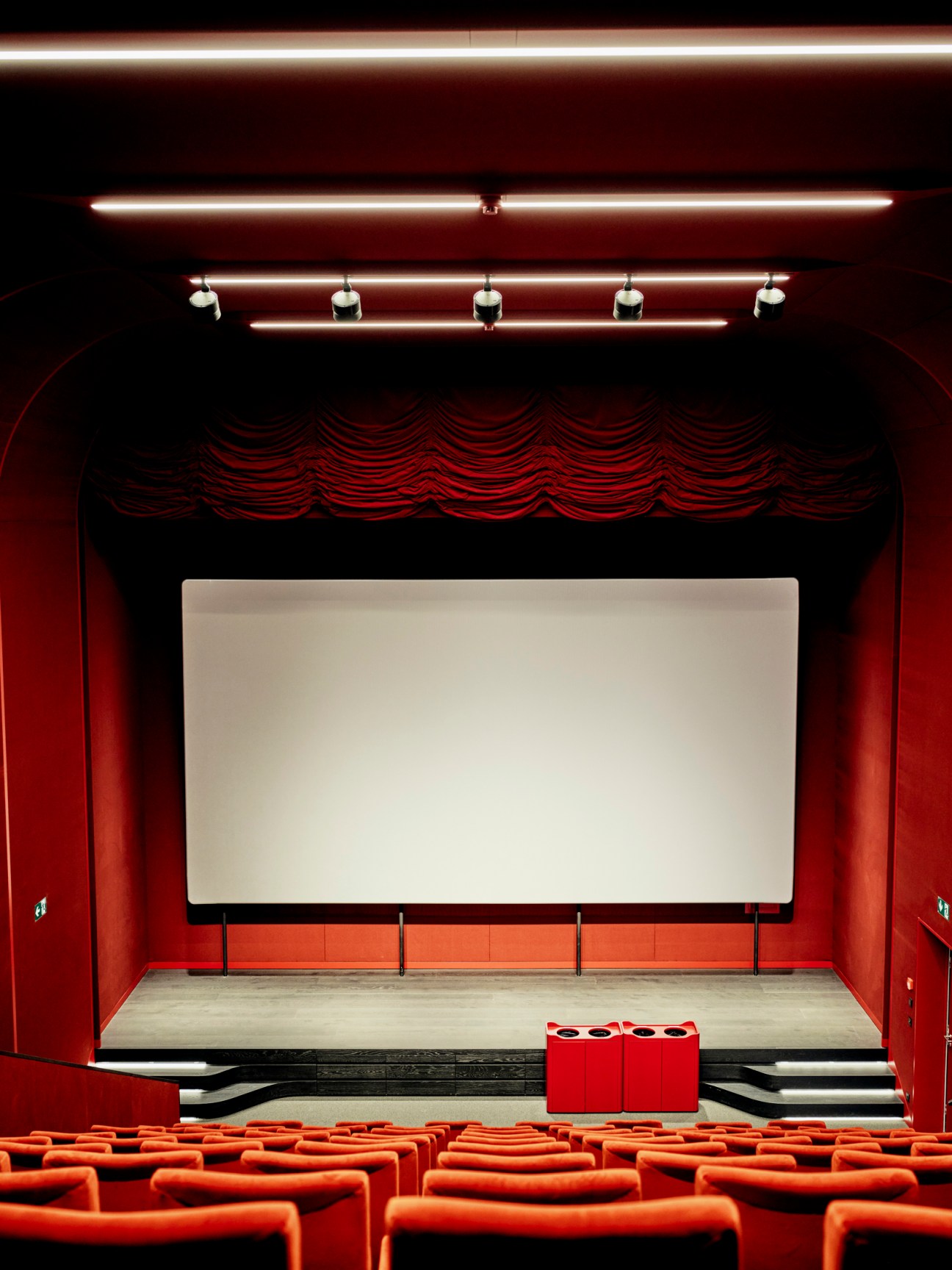 Red cinema auditorium with rows of red seats facing a white screen and stage