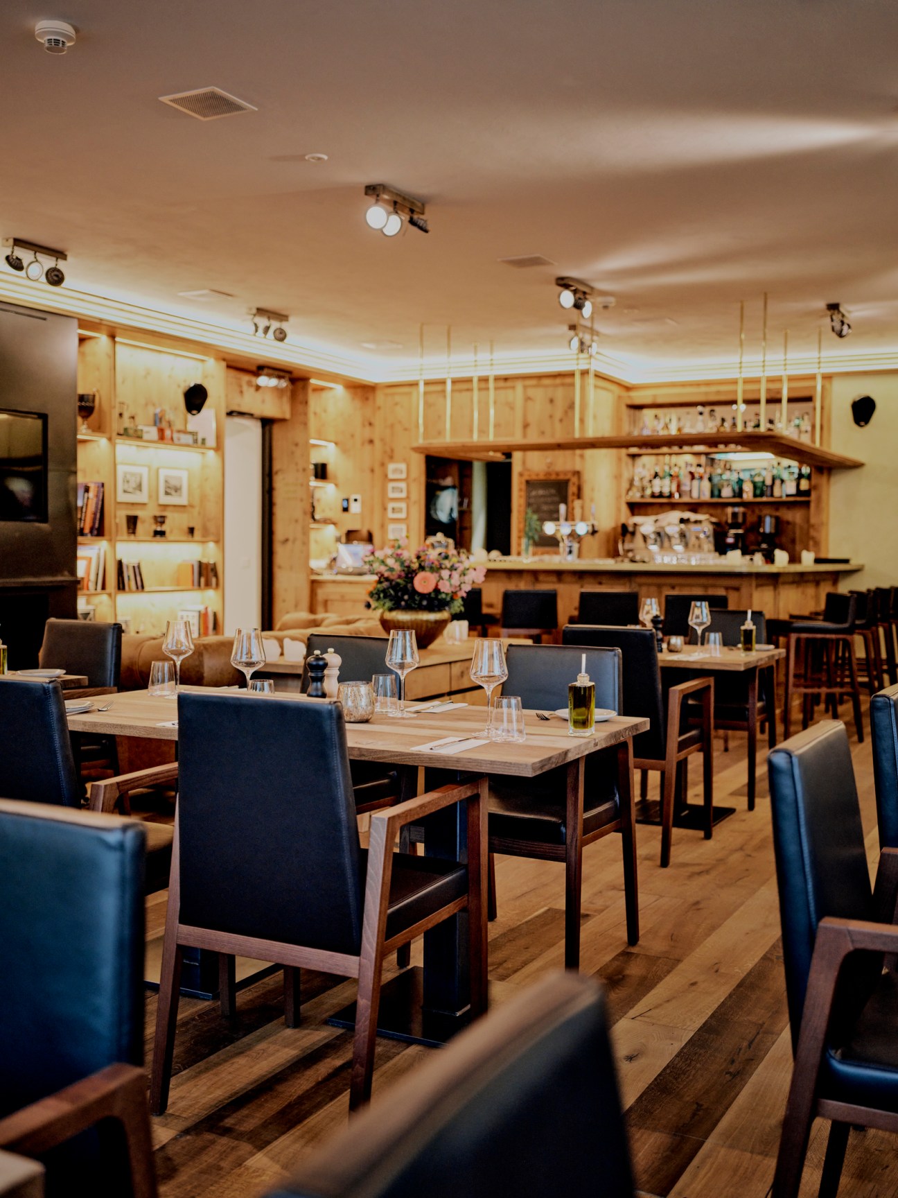Restaurant interior with wooden walls, dark chairs at set tables, and bar in background