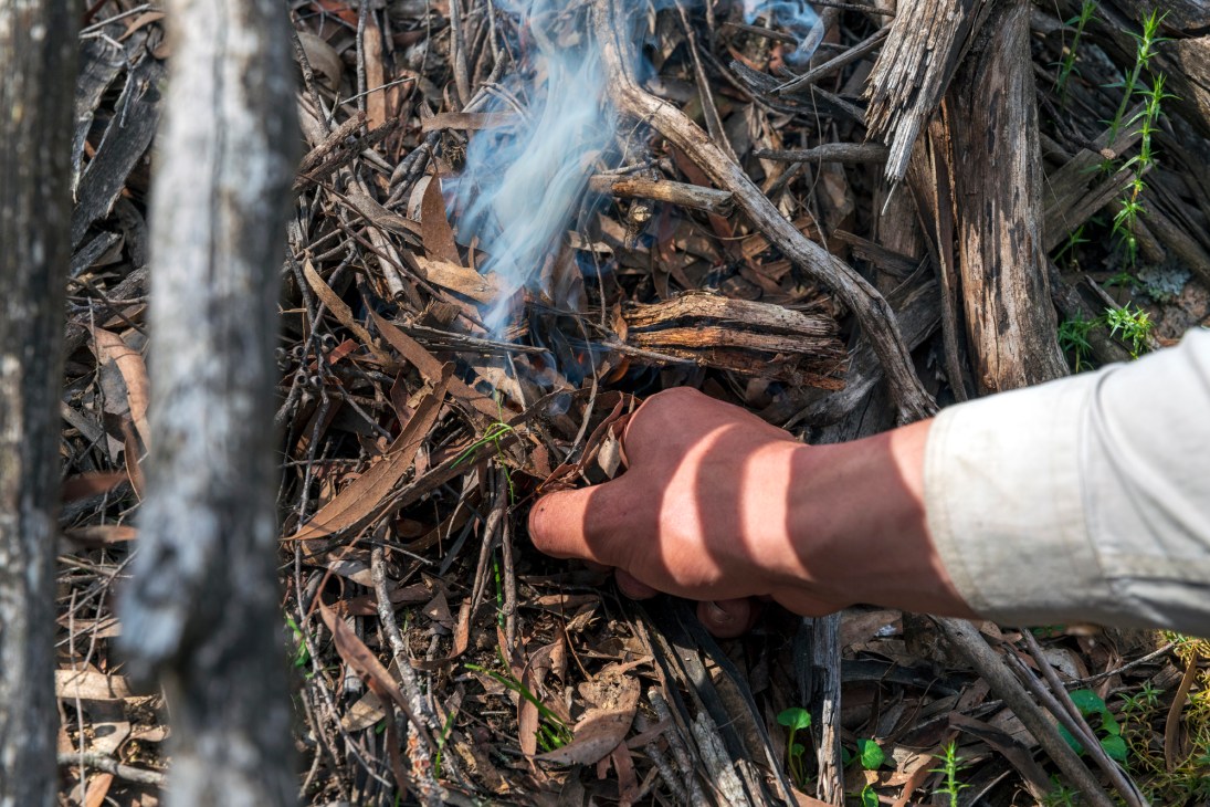A person's hand guides a controlled burn among dry twigs and charred wood in the Australian bush.