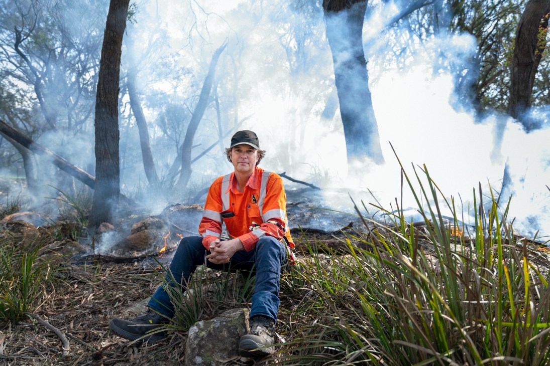 Indigenous-burning practitioner Den Barber crouches in the Australian bush during a controlled burn, wearing orange fire-resistant clothing.