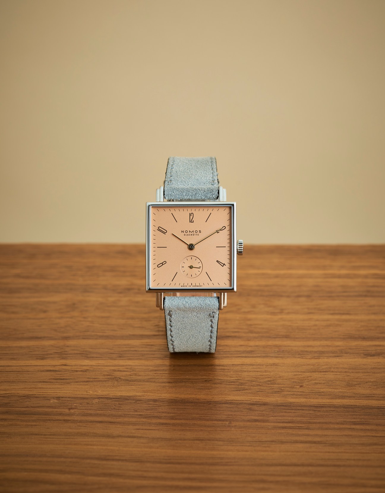 Square-faced Nomos Glashütte Tetra Divine Spark watch with salmon-colored dial and gray leather strap on wood surface
