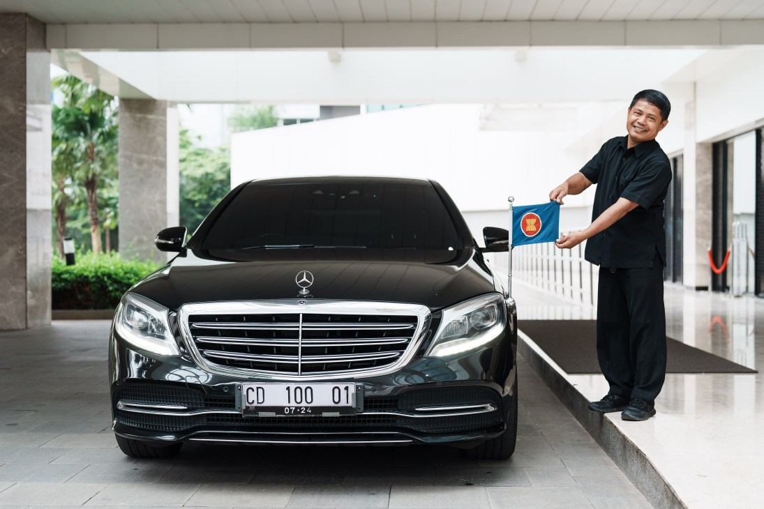 A smiling man in dark clothing stands beside a black Mercedes-Benz sedan holding a small ASEAN flag