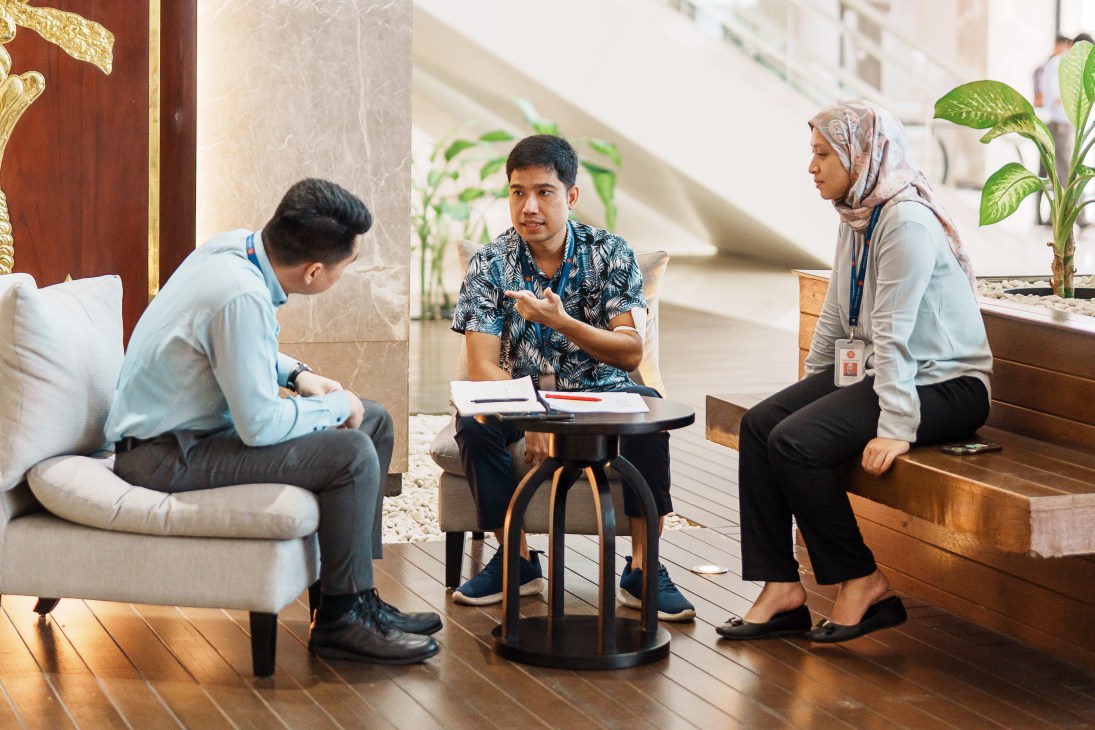 Three ASEAN staff members having a discussion in a modern lobby with wooden floors and marble columns