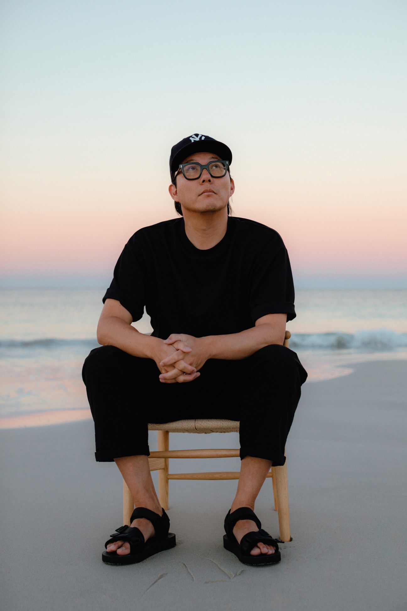 Ken Leung sits on a wooden stool on a beach at sunset wearing all black clothing and a baseball cap