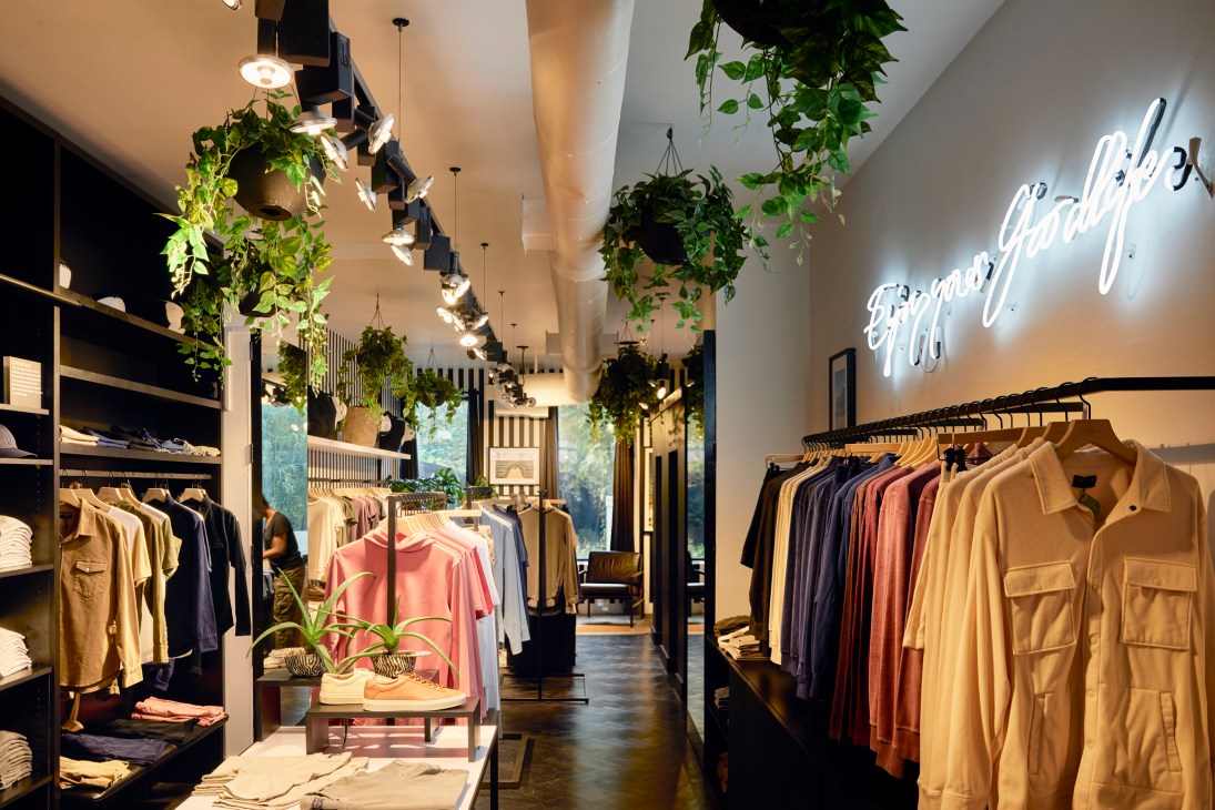 Interior of a modern retail boutique with hanging plants, neon signage, and clothing displayed on racks and shelves.