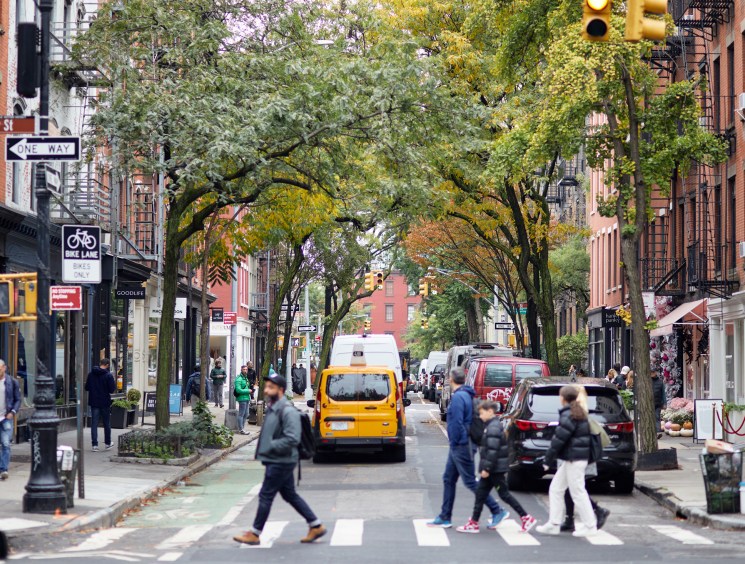 Tree-lined street in Greenwich Village with pedestrians crossing, storefronts, yellow taxi and parked cars on Bleecker Street.