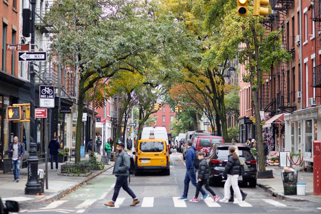 Tree-lined street in Greenwich Village with pedestrians crossing, storefronts, yellow taxi and parked cars on Bleecker Street.