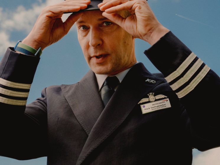Mark Vanhoenacker in British Airways pilot uniform adjusting his cap against a cloudy sky