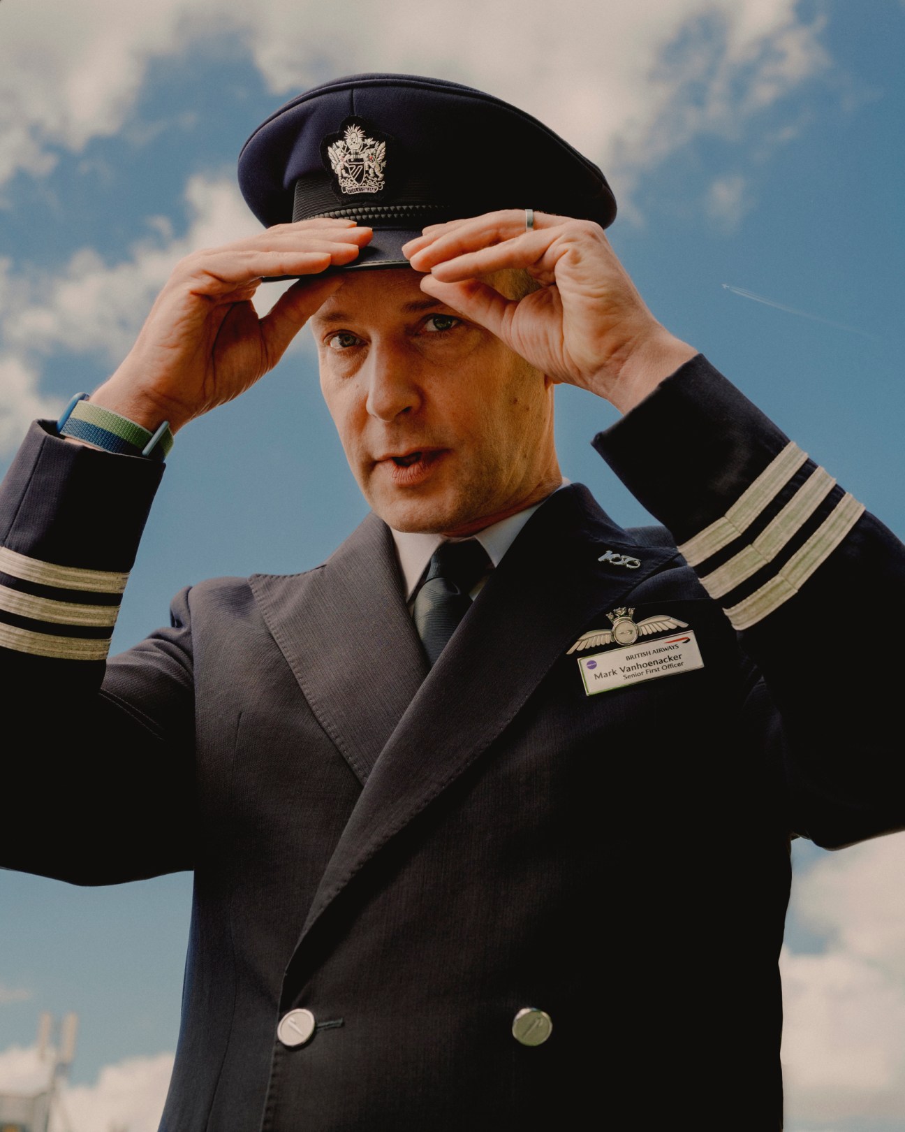 Mark Vanhoenacker in British Airways pilot uniform adjusting his cap against a cloudy sky