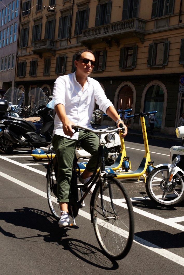 Man in white shirt and sunglasses riding black bicycle on Milan street with yellow scooters parked nearby