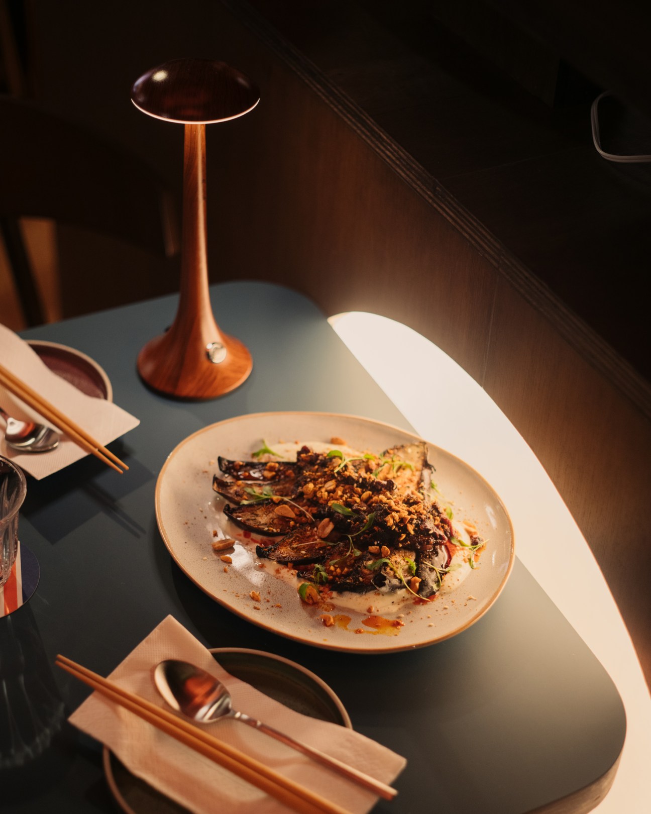 A plate of charred aubergine with garnishes on a dark restaurant table with wooden chopsticks and ambient lighting