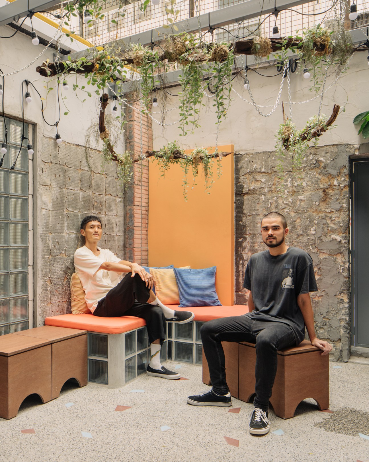 Two men sitting on orange cushioned benches in an industrial-style interior with exposed brick walls and hanging plants