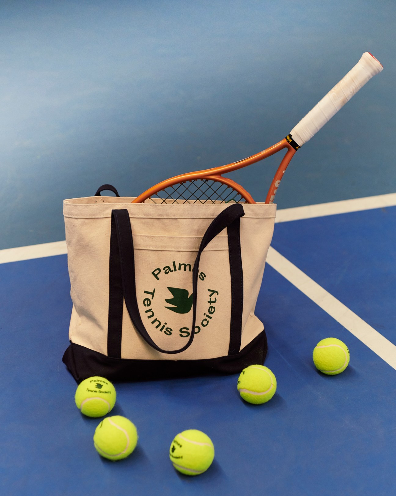 Palmes Tennis Society canvas tote bag with tennis racket and yellow tennis balls on blue court surface