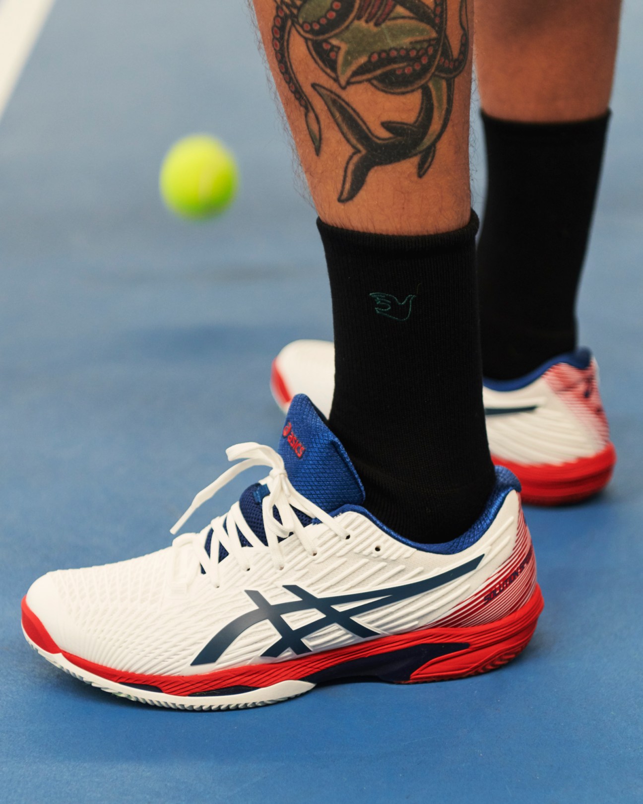 Close-up of tennis player's feet wearing white Asics shoes and black Palmes socks on blue court