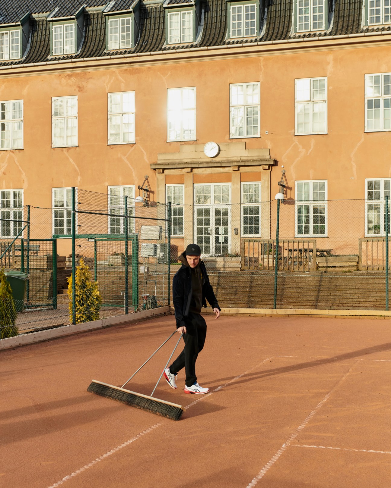 Person in black athletic wear using a large brush to sweep lines on a red tennis court surface