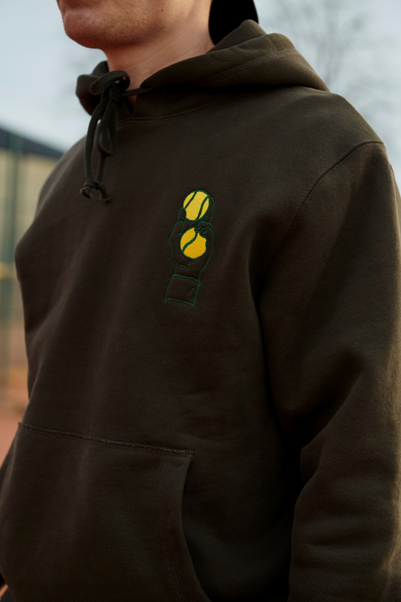 Person wearing olive green hooded sweatshirt with yellow tennis ball embroidered logo