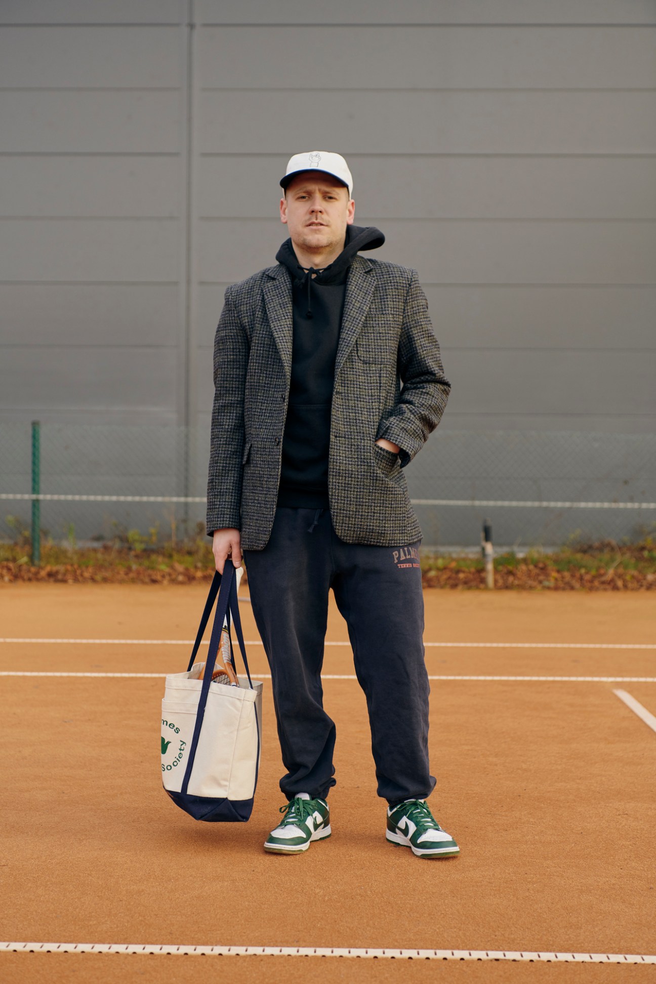 Nikolaj Hansson wearing a gray blazer over navy hoodie and sweatpants, holding a tote bag on a tennis court