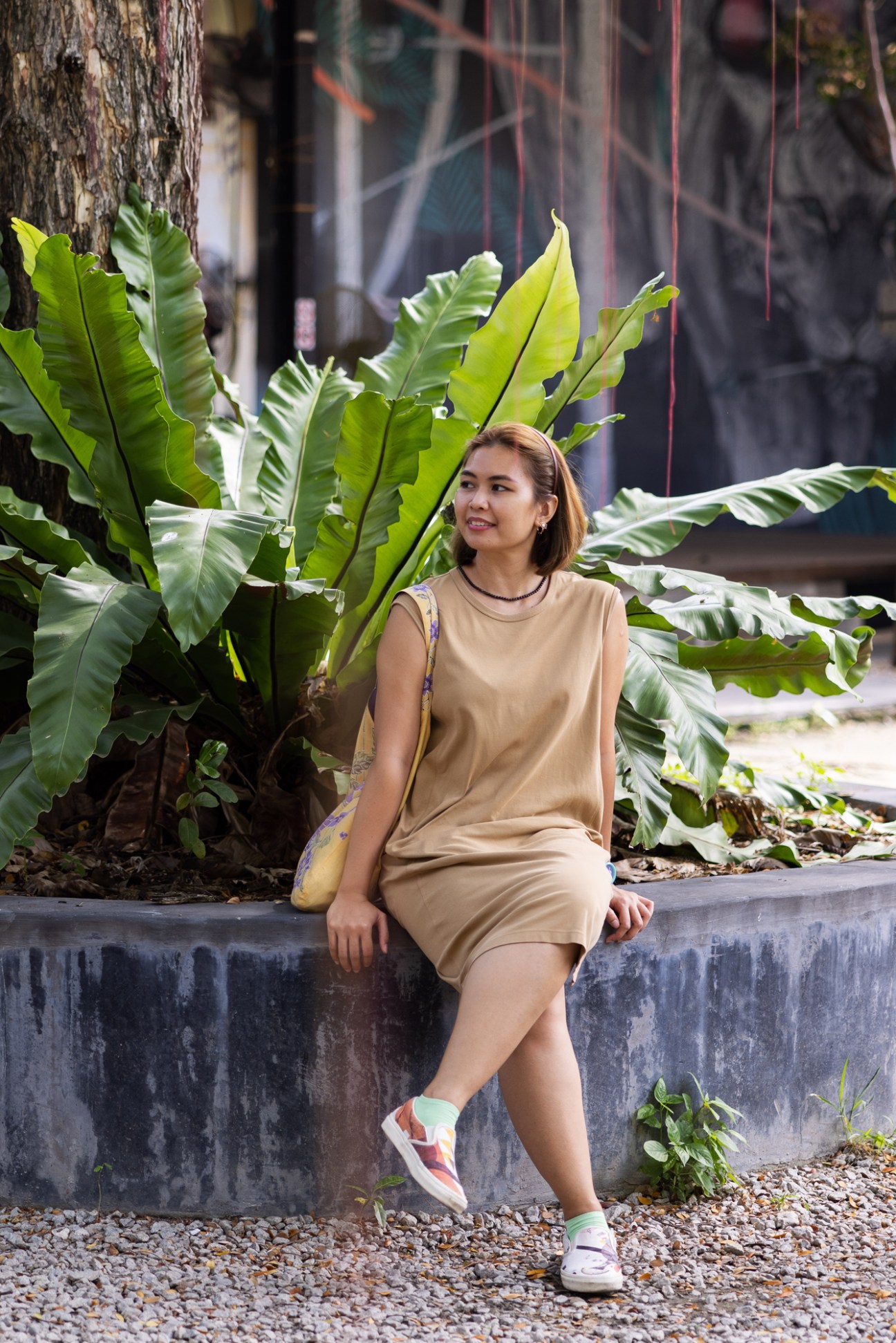 Woman in beige dress sitting on concrete ledge surrounded by large tropical plants in outdoor setting