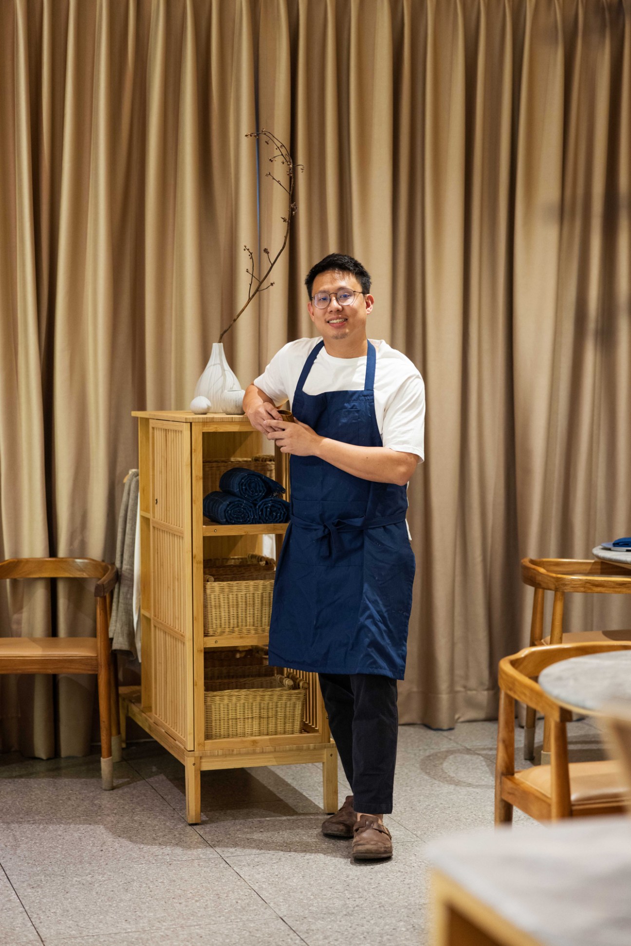 Johnson Wong wearing blue apron and glasses standing next to wooden shelving unit in restaurant interior