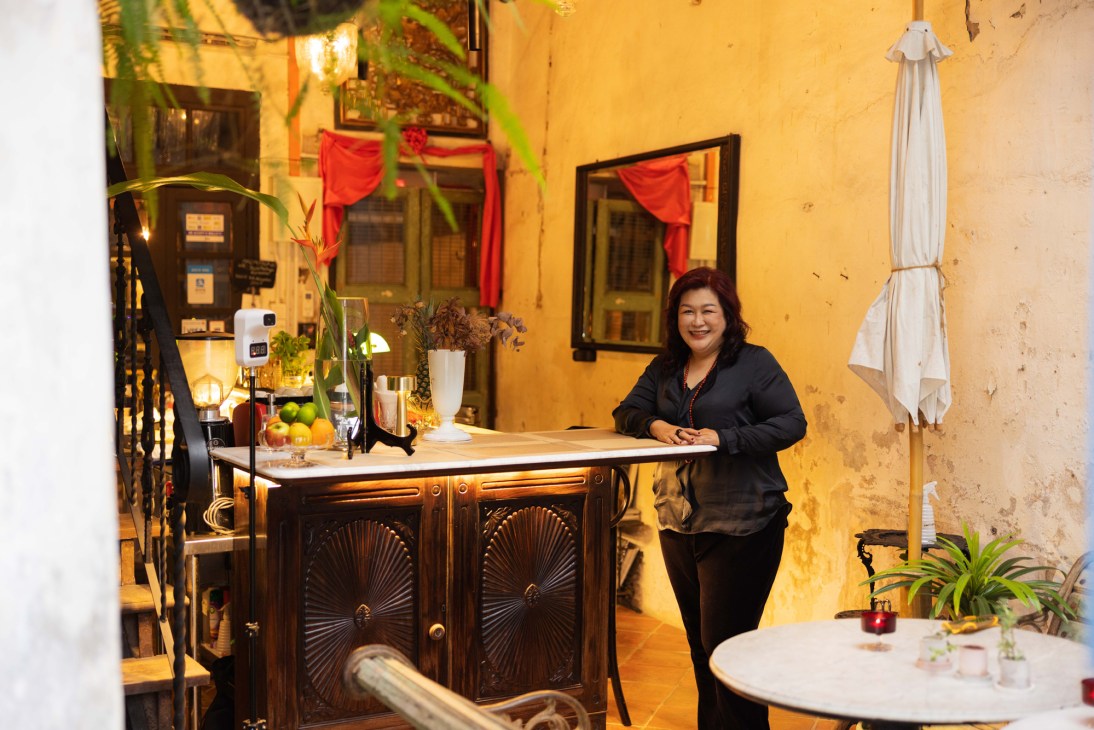 Woman in black outfit standing behind ornate wooden bar counter in atmospheric courtyard setting