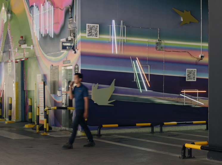 Person walking past large colorful mural in industrial warehouse space with barriers and equipment visible