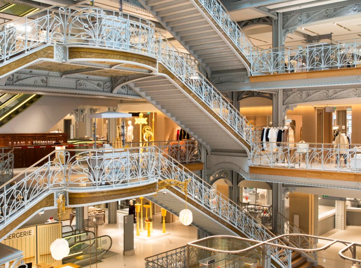 Multi-level department store interior with ornate white cast iron railings, curved staircases, and luxury retail displays