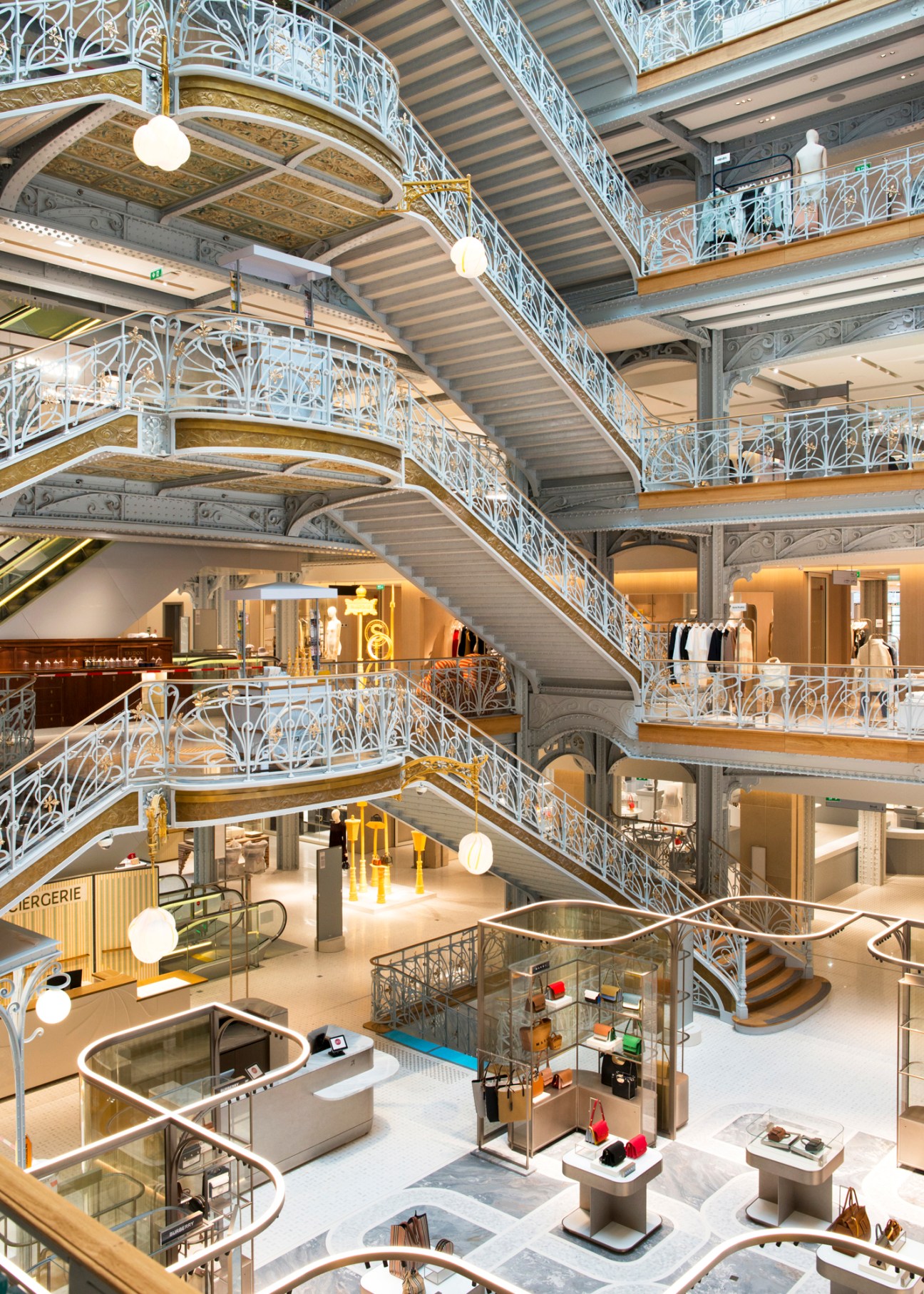Multi-level department store interior with ornate white cast iron railings, curved staircases, and luxury retail displays