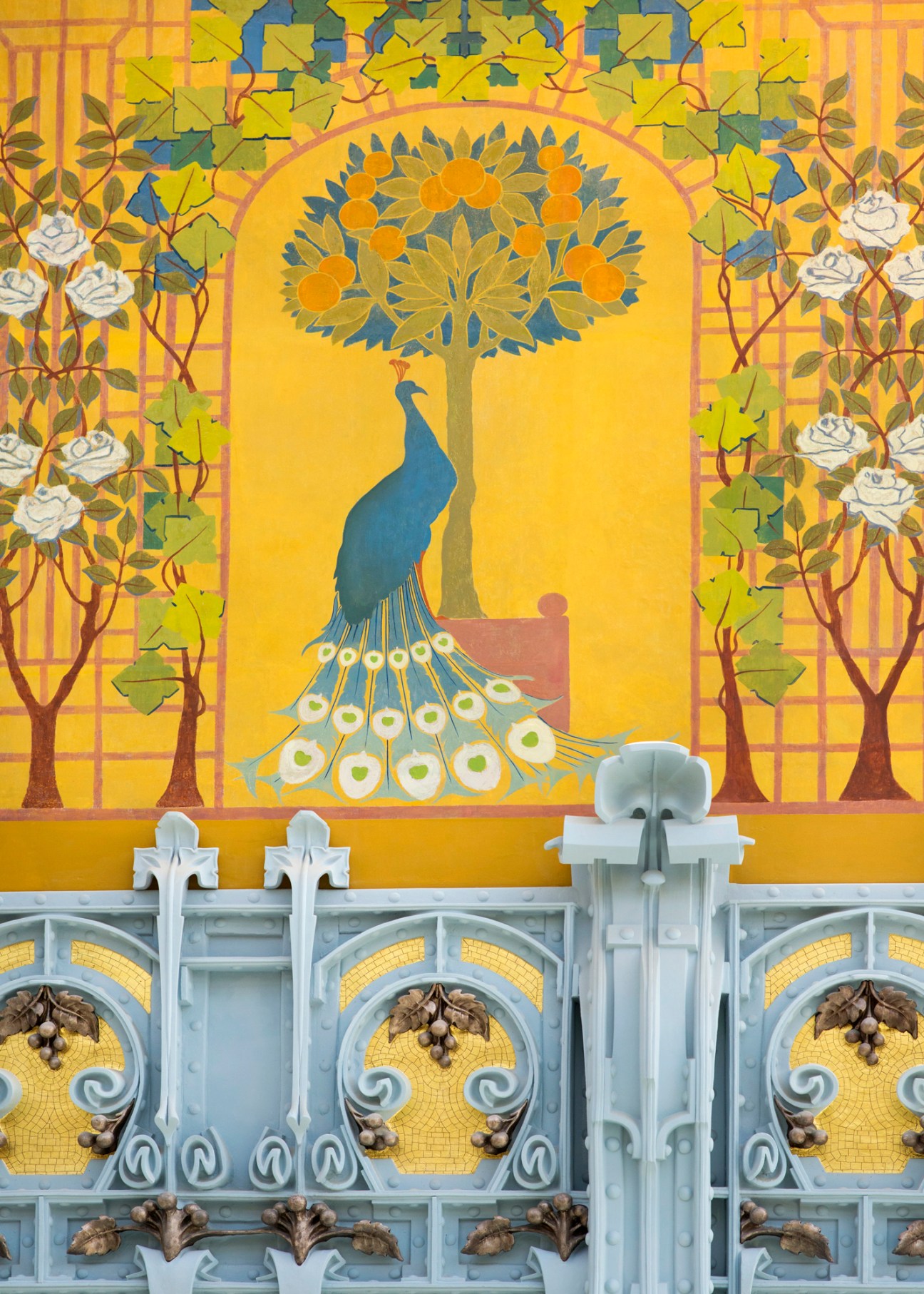 Ornate Art Nouveau mural featuring a blue peacock beneath a fruit tree, surrounded by rose vines and decorative ironwork