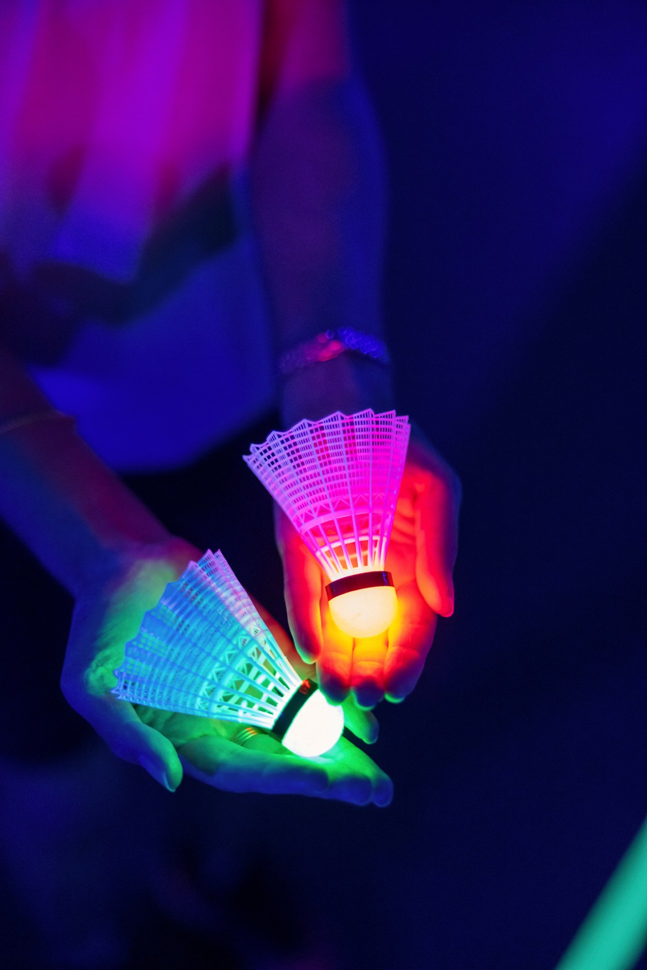 Two hands holding glowing badminton shuttlecocks under neon lighting in darkness