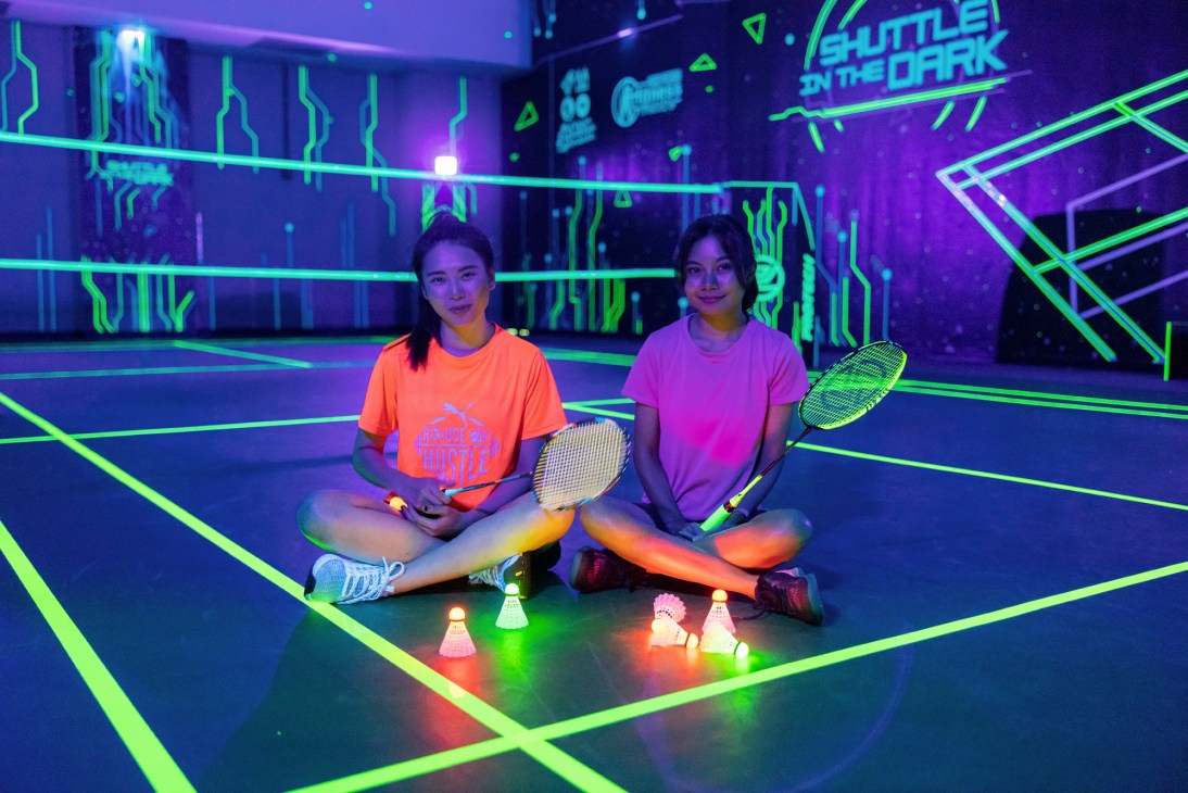Two women sitting cross-legged on a glow-in-the-dark badminton court holding rackets with illuminated shuttlecocks