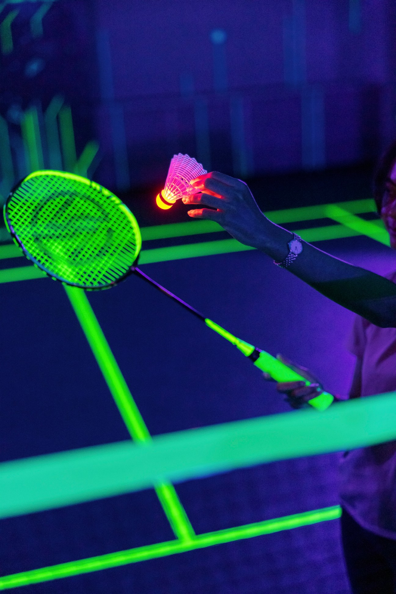 Hand holding glowing shuttlecock above illuminated badminton net in darkened court with neon lighting