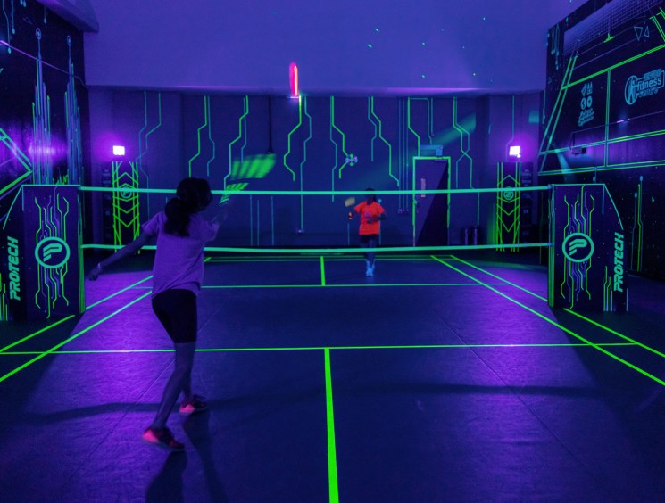 Players on a glow-in-the-dark badminton court with neon green lines and purple lighting at Shuttle in the Dark