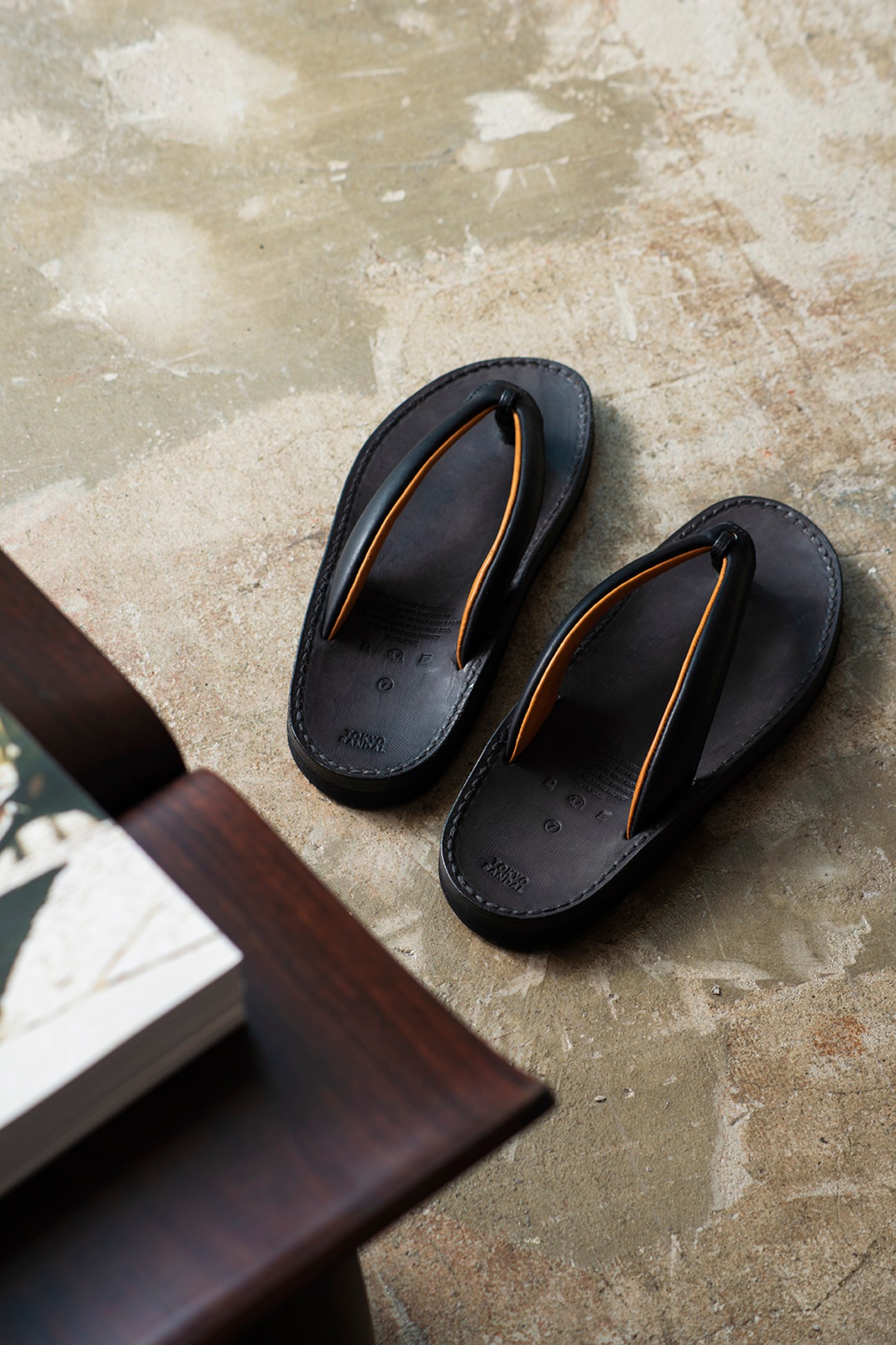 Pair of black leather setta sandals with orange straps on sandy concrete surface