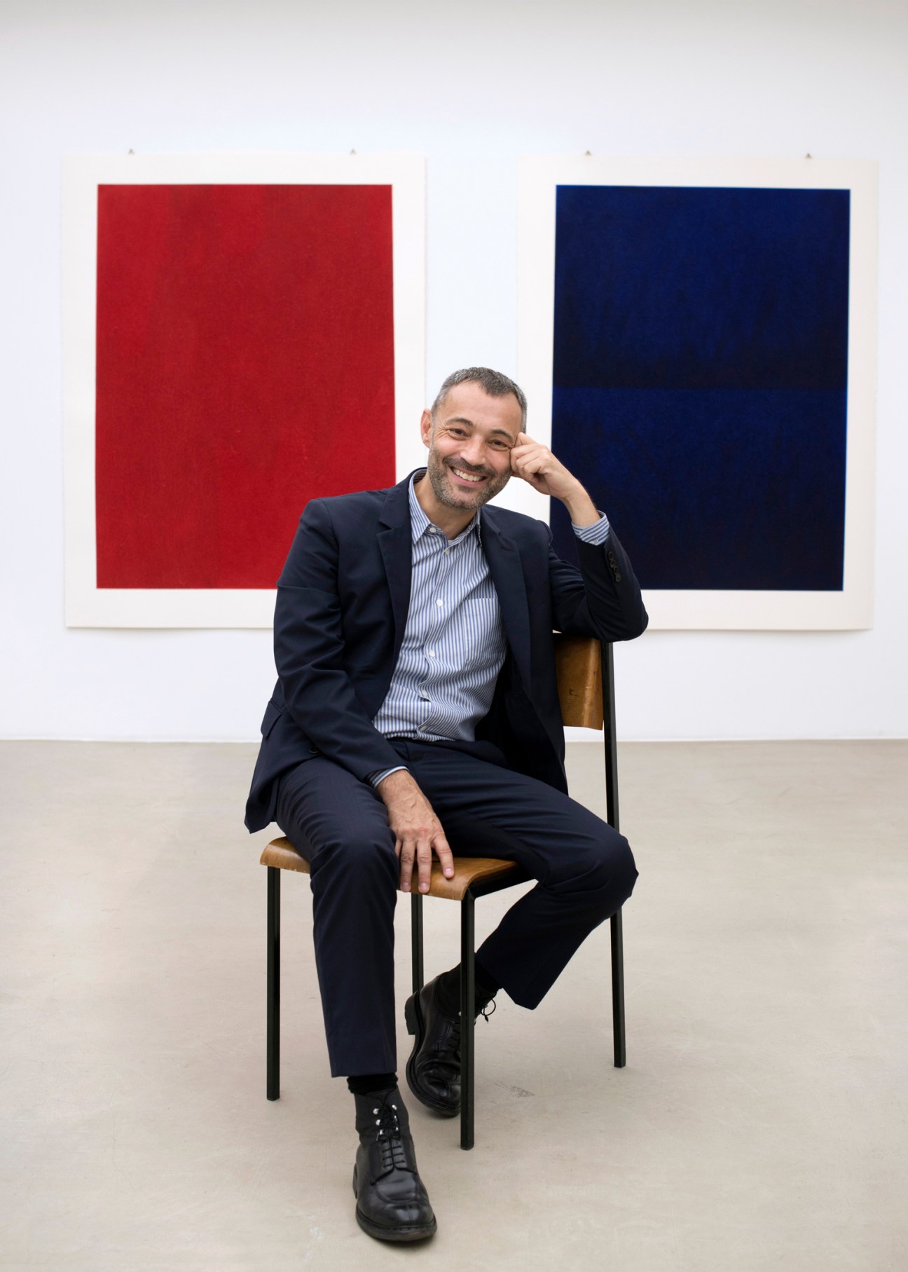 Gallerist Jérôme Poggi smiling in his Paris gallery seated in front of red and blue artworks