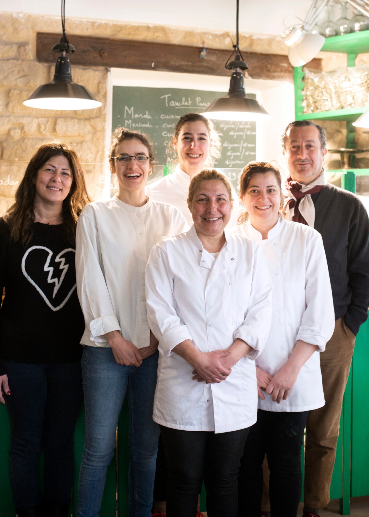 Group of six restaurant staff members posing together in kitchen with stone walls and pendant lighting