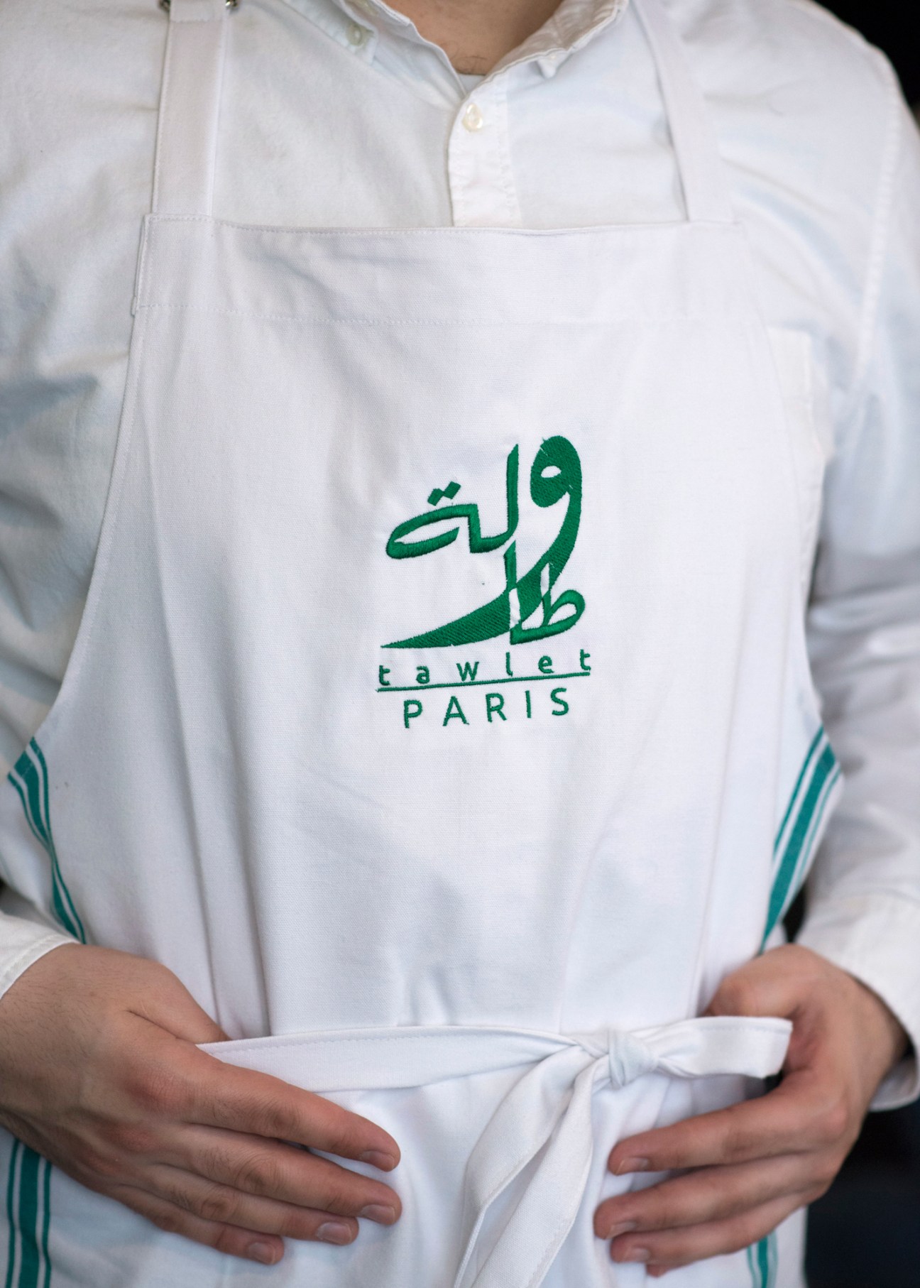 White apron with green Arabic text and 'tawlet PARIS' branding hanging in restaurant