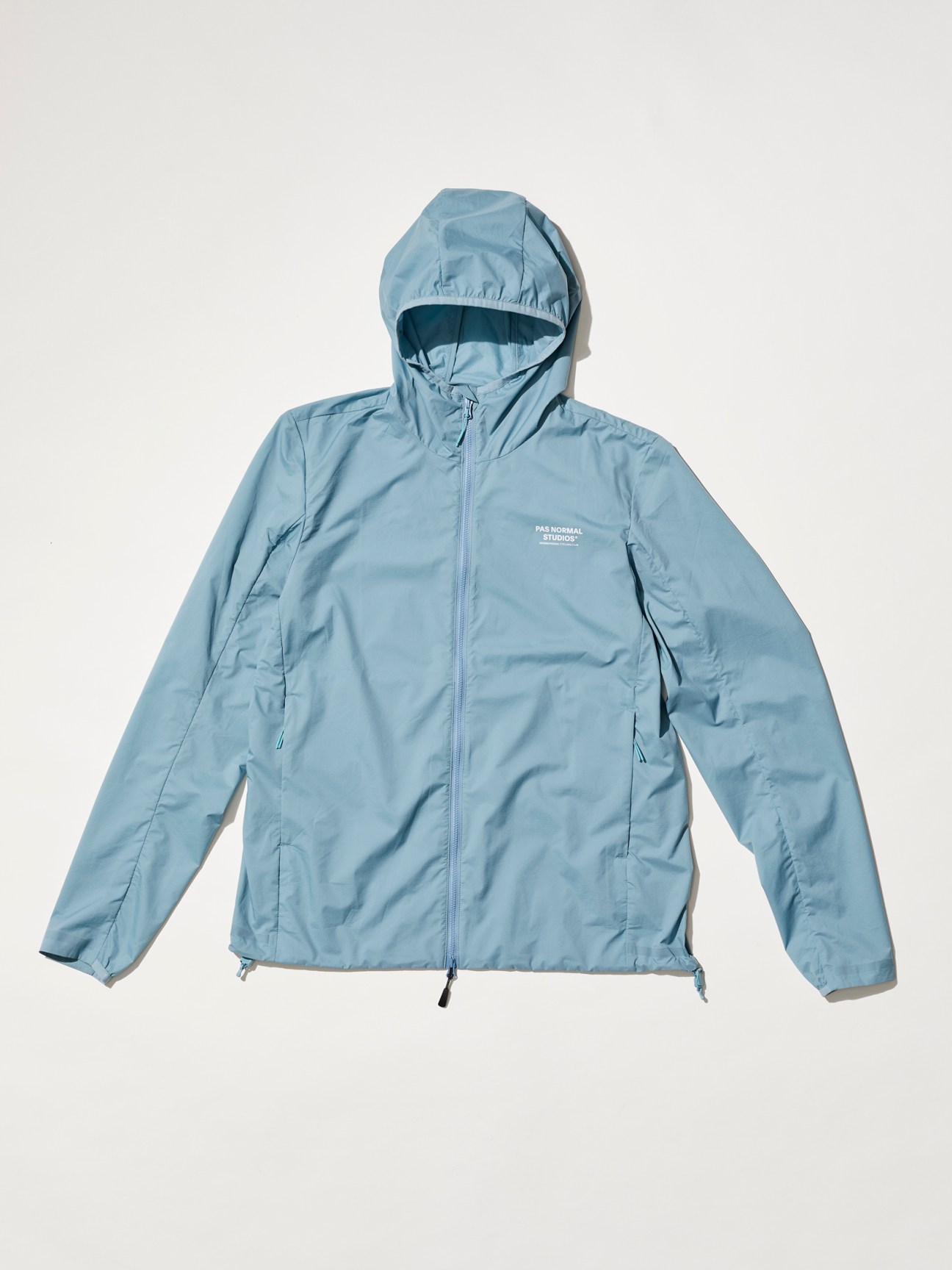 Light blue hooded cycling jacket with full zip closure and brand logo on chest which is a Pas Normal Studios dusty blue ripstop cycling jacket with integrated storage pocket