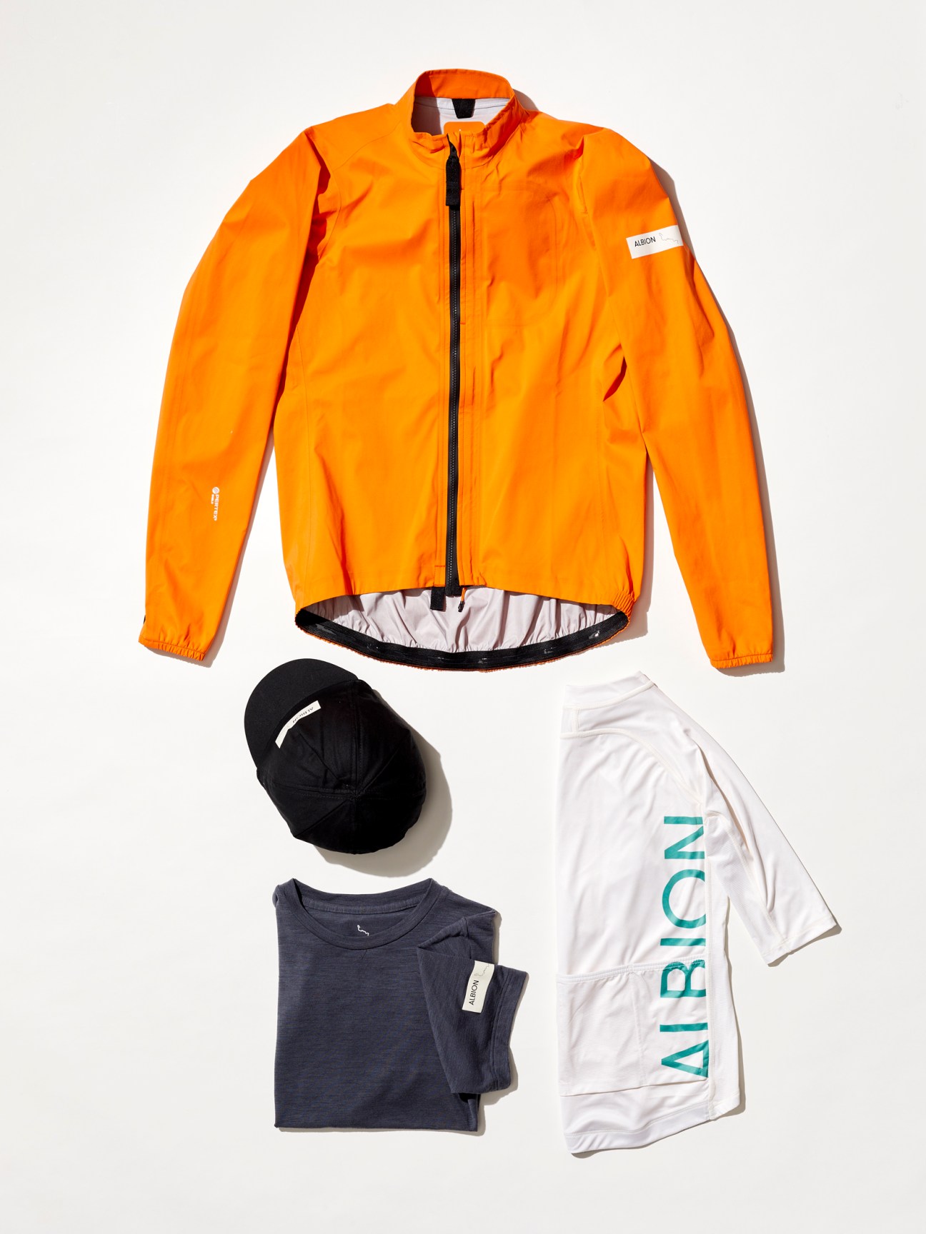 Albion cycling gear Orange cycling jacket with reflective lining displayed with black cap, navy t-shirt and white Albion branded shirt