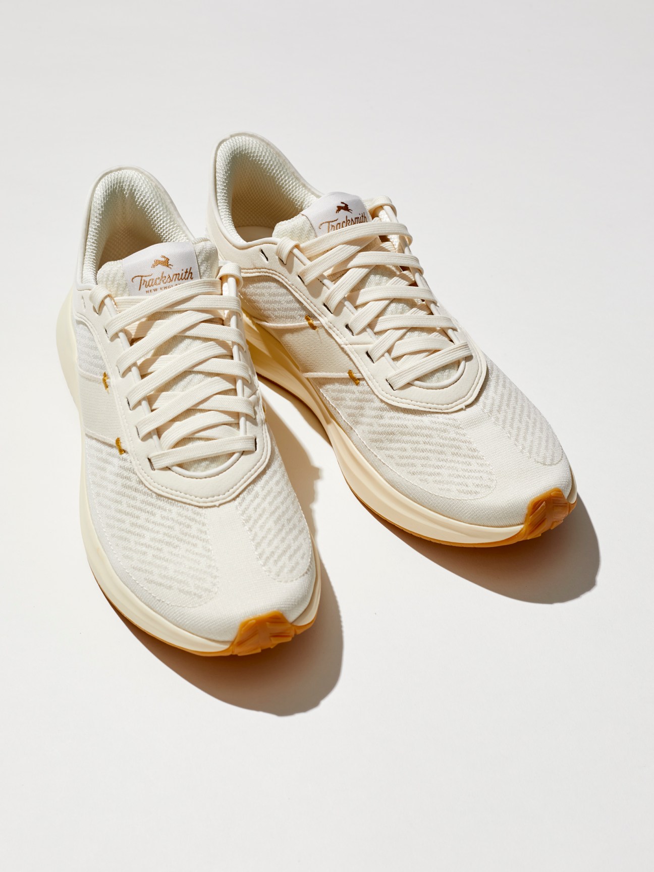 Pair of cream-colored Tracksmith Eliot Runner sneakers with woven mesh pattern and orange sole accents on white background