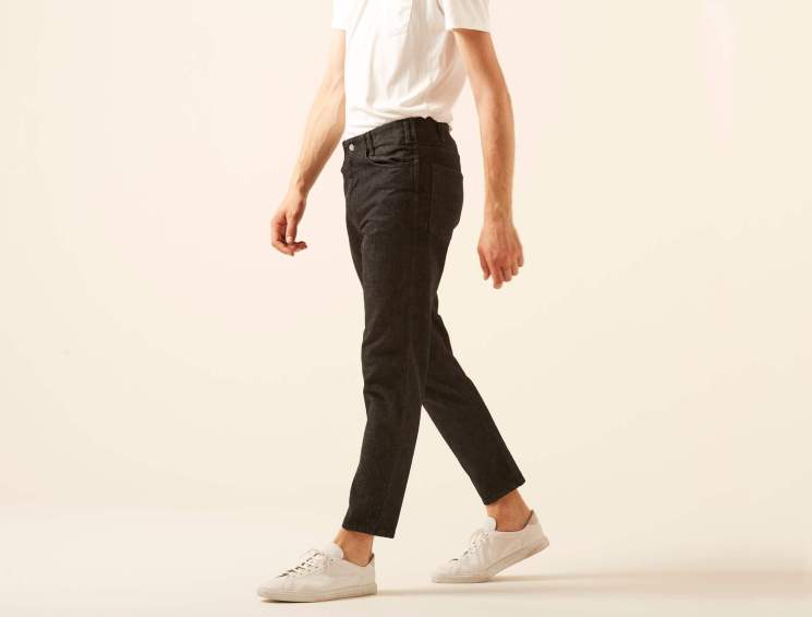 Person in white shirt and dark jeans walking, wearing white sneakers against neutral background