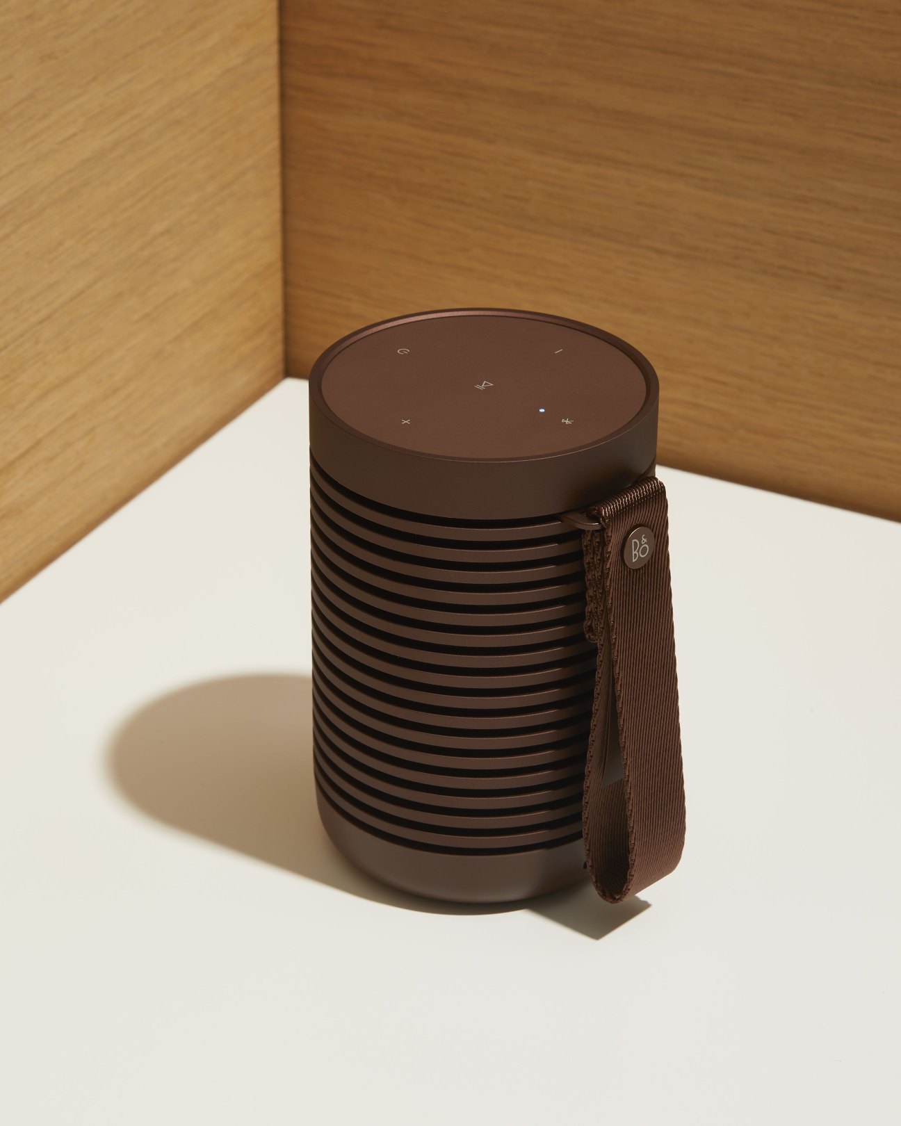 Brown cylindrical Bang & Olufsen Beosound Explore speaker with fabric strap and touch controls on white surface