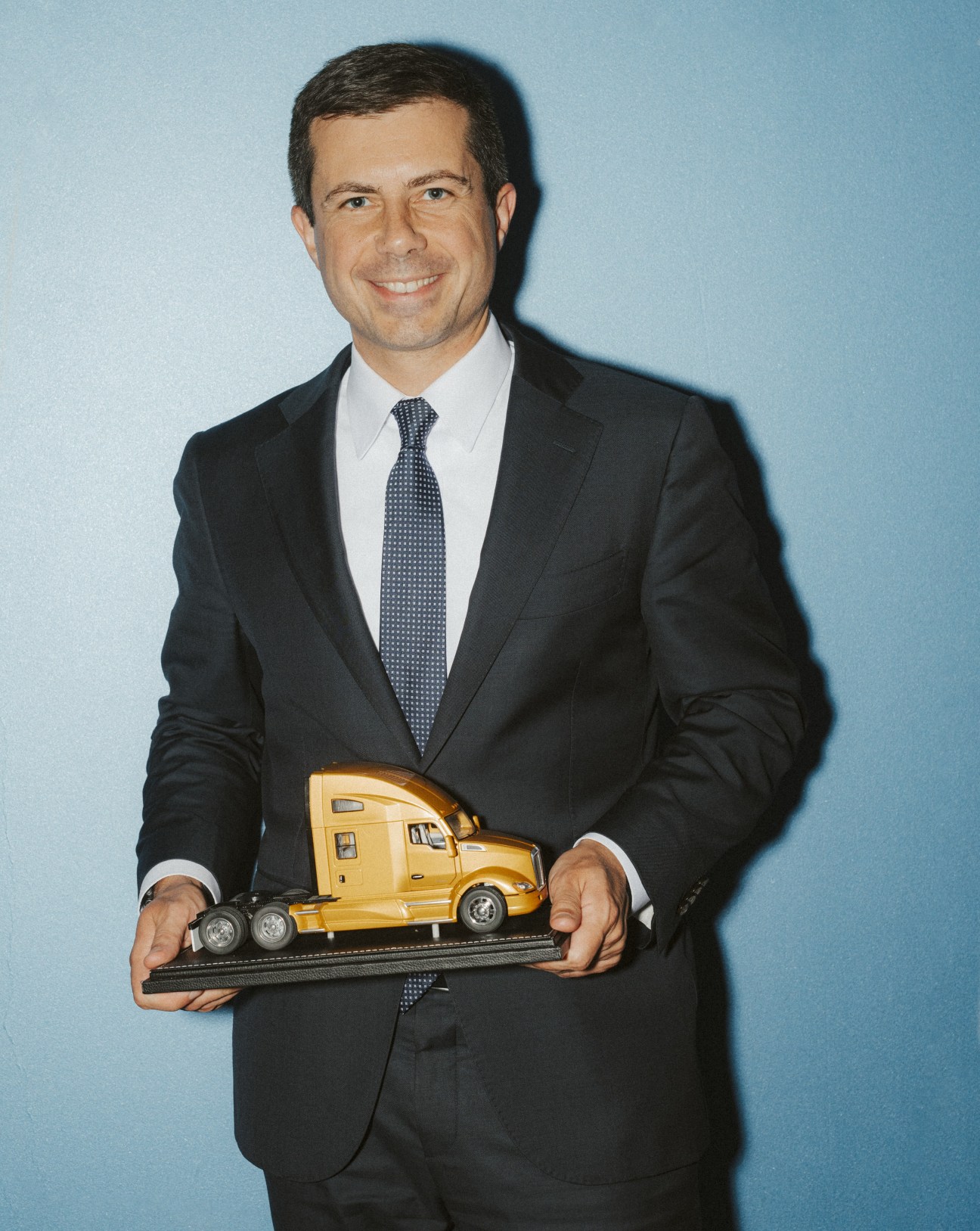 Pete Buttigieg in dark suit and blue tie holding a toy yellow truck against a light blue background