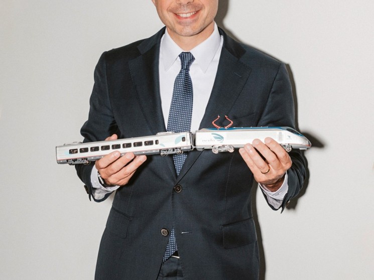 Pete Buttigieg in dark suit and blue tie holding a model passenger train against white background