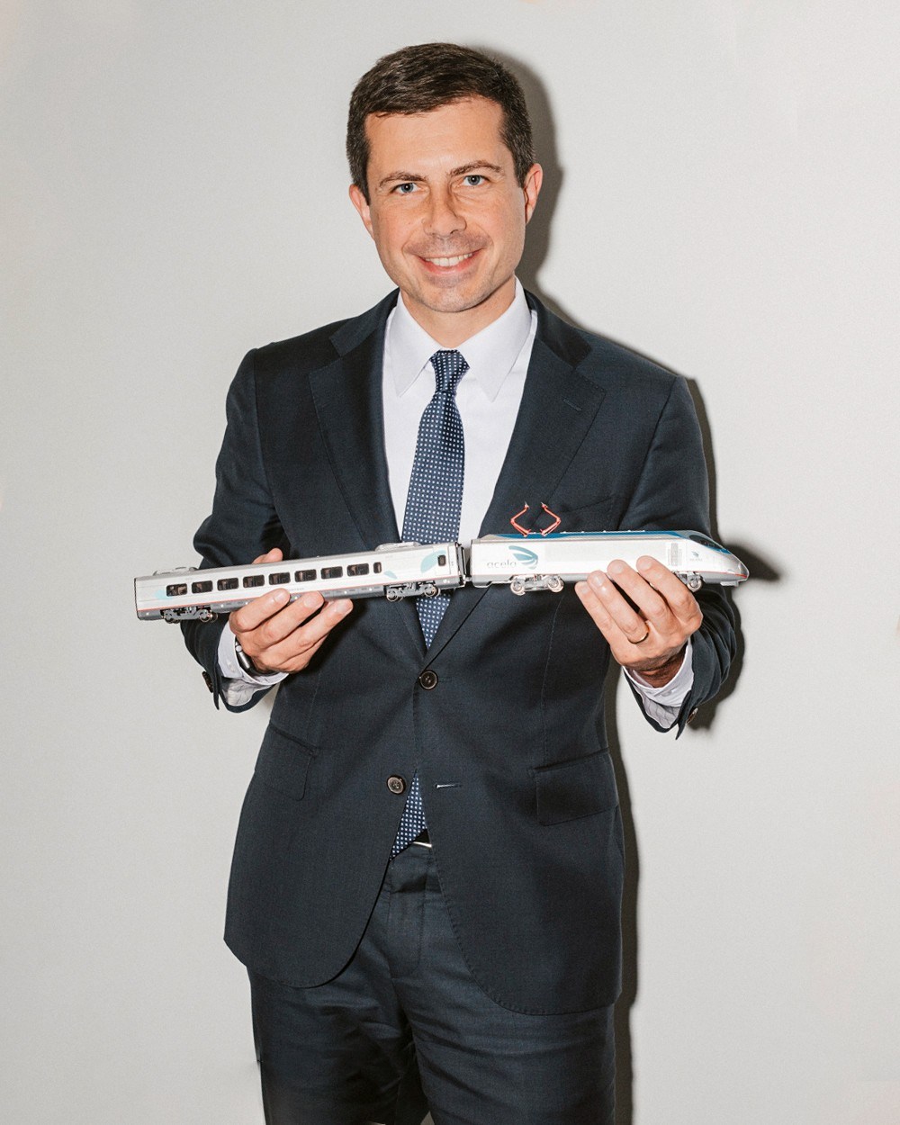 Pete Buttigieg in dark suit and blue tie holding a model passenger train against white background