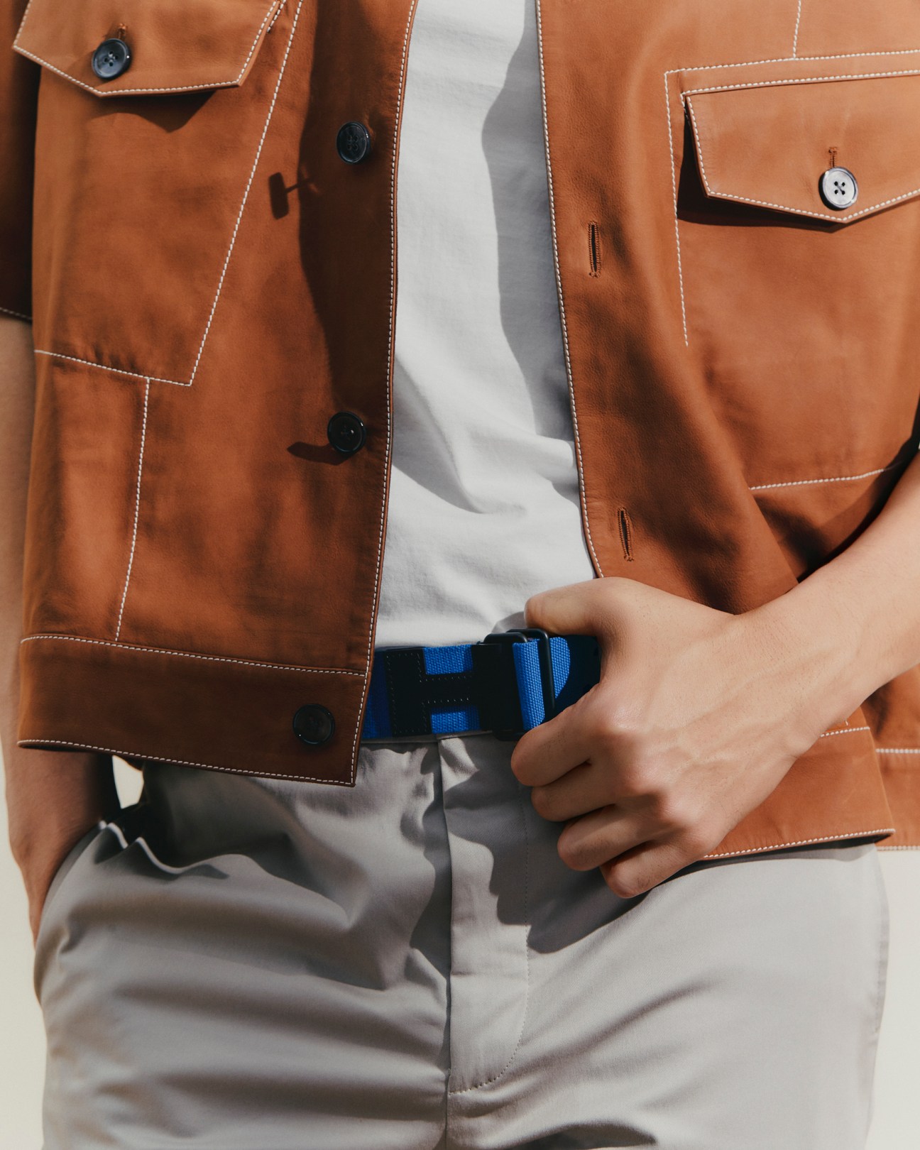 Man wearing brown suede jacket with white contrast stitching and blue webbed belt, hand adjusting belt buckle