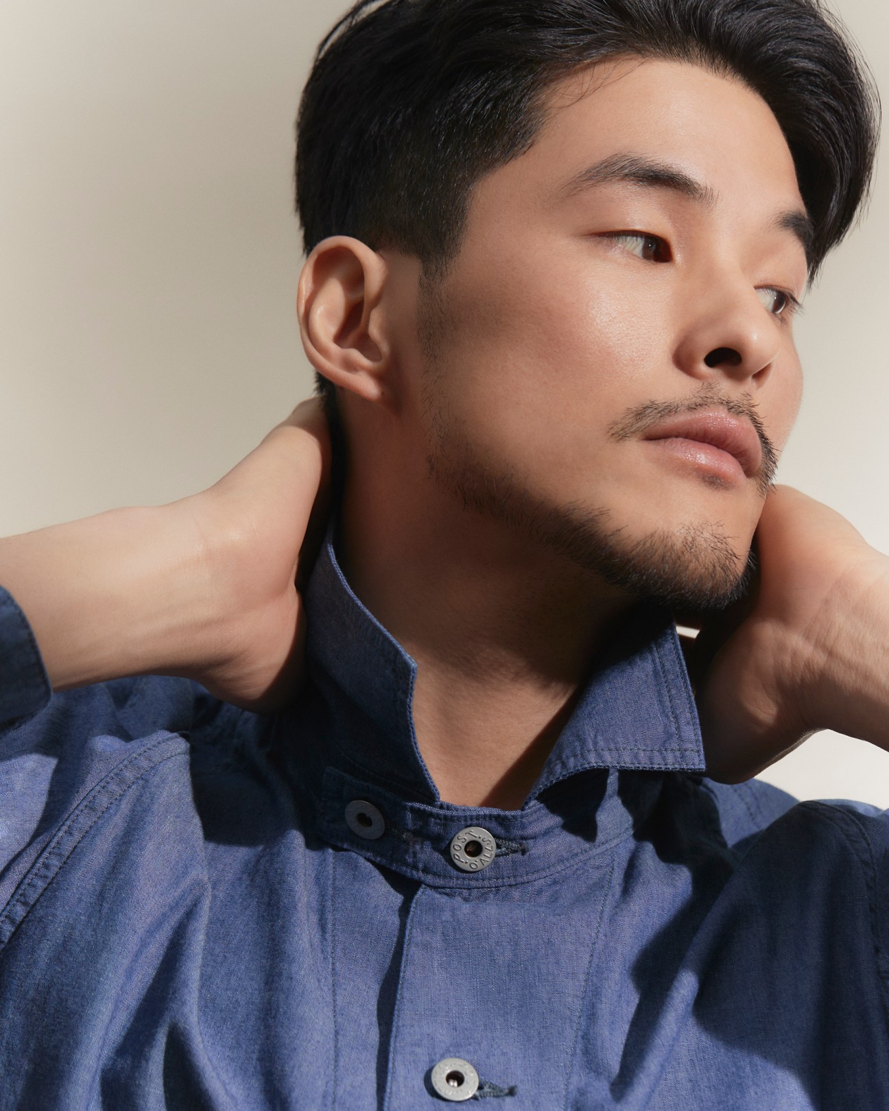 Asian man in blue denim shirt with hands behind neck looking to the side in contemplative pose