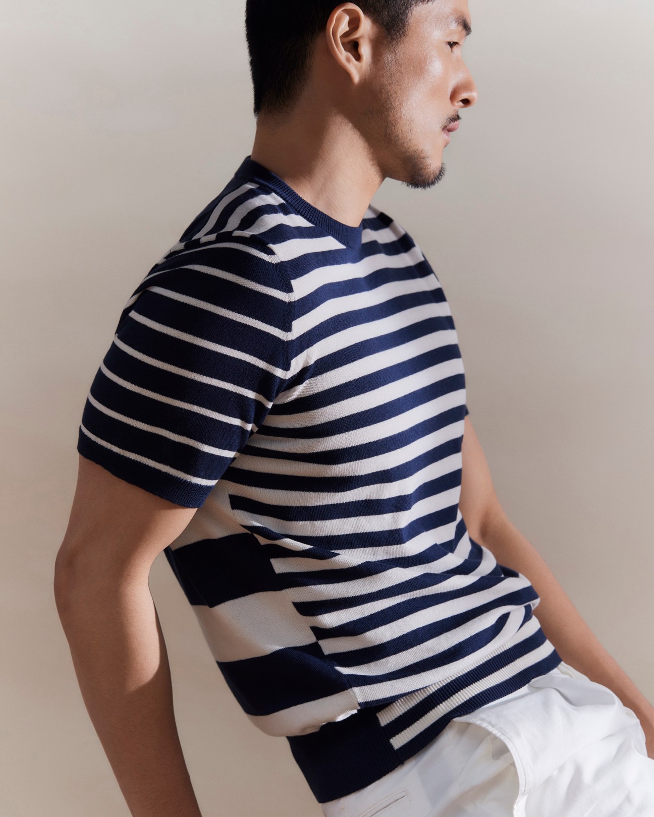 Man in profile wearing navy and white striped knit sweater with white trousers against neutral background