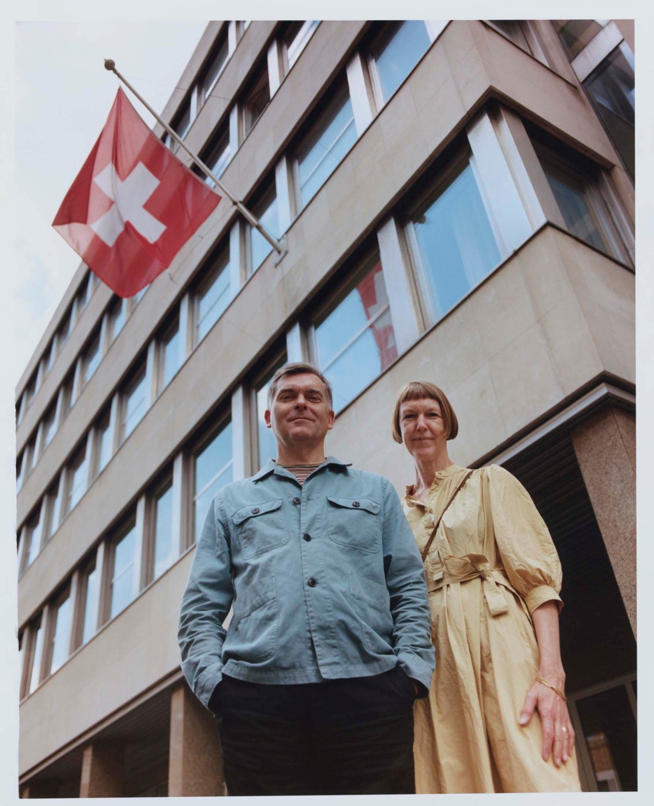 Two people standing outside a modernist building with a Swiss flag overhead.