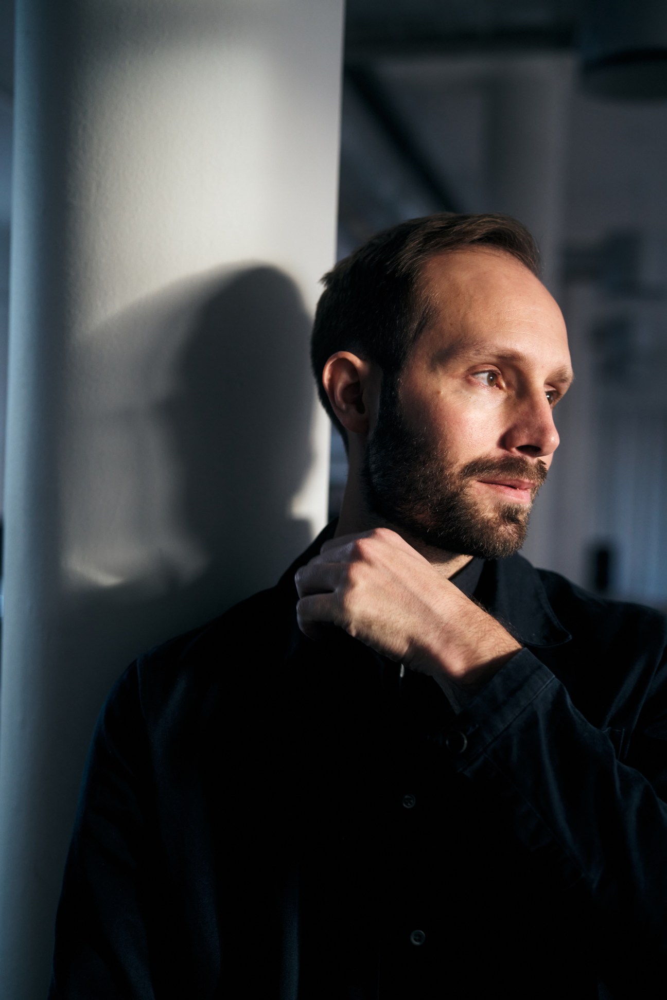 Portrait of a bearded man in a dark shirt against a minimalist white wall with blue vertical element.