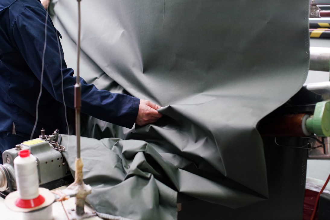 Person in blue jacket using steam iron on dark waxed cotton fabric in textile manufacturing facility
